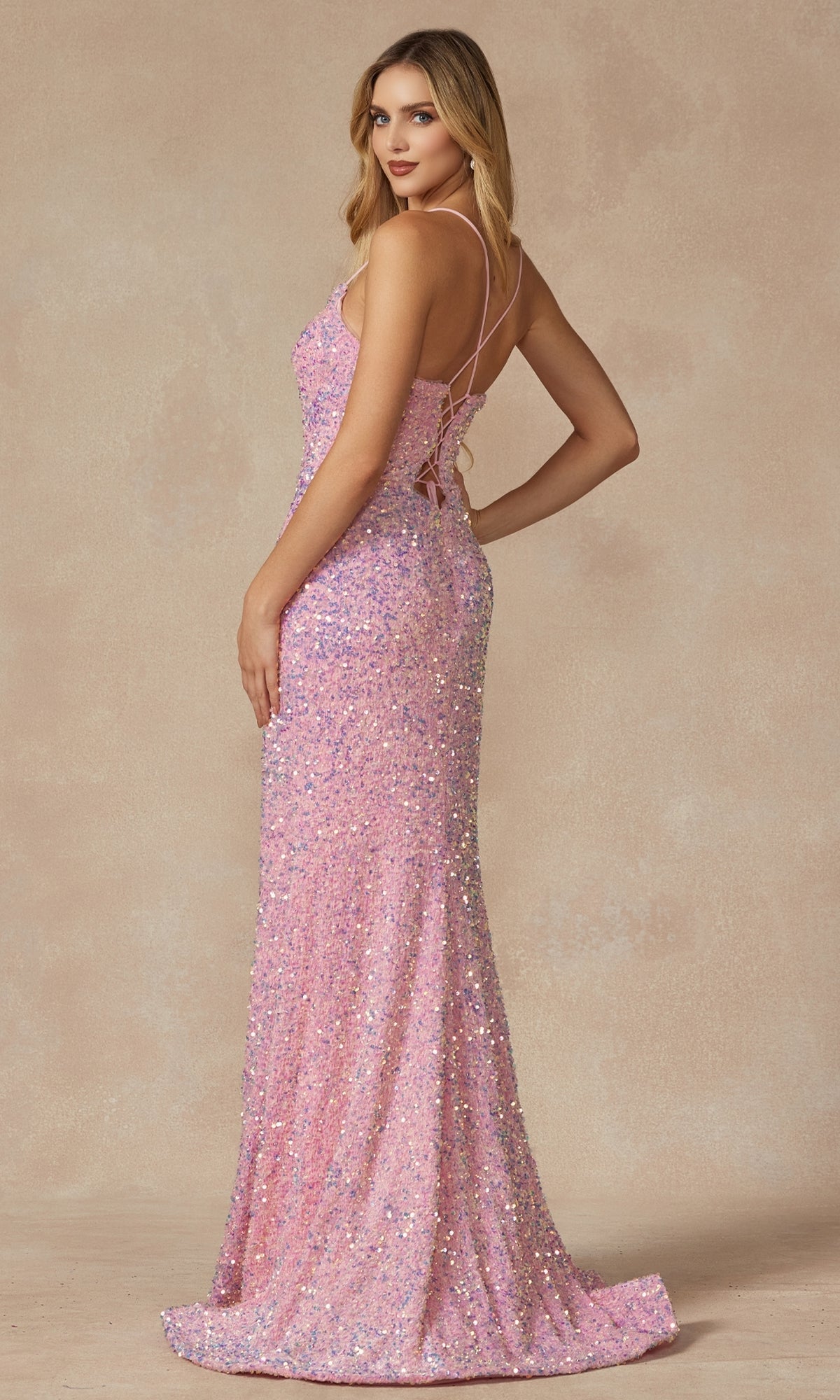 A woman models the V-Neck Long Sequin Prom Dress 2406, a pink floor-length gown with thin straps and an open lace-up back, standing on a neutral background facing slightly away from the camera.